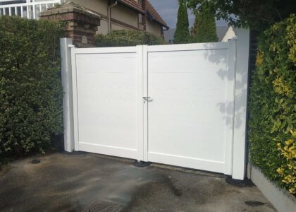 Portail battant blanc fermé entre des haies verdoyantes.